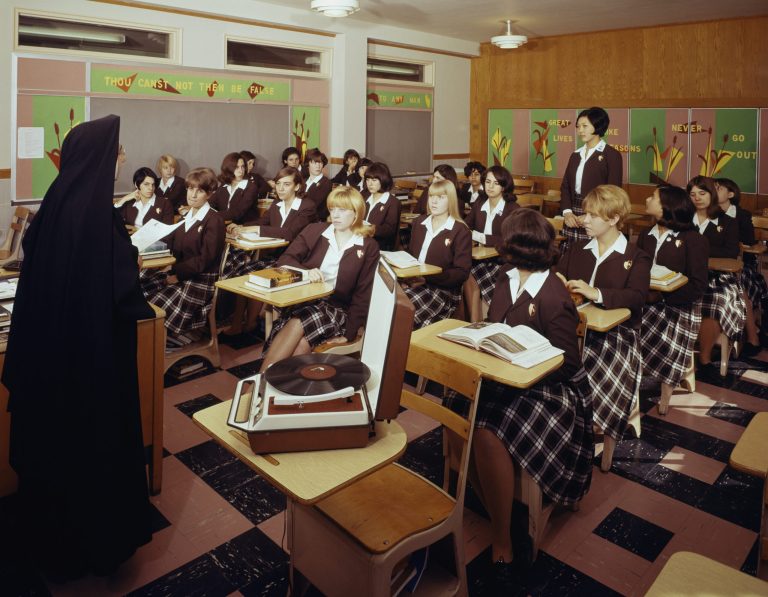 Nun teaching class of girls in uniforms with books.