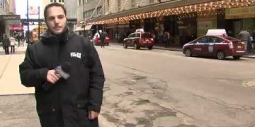 Man reporting on city street with taxis passing.