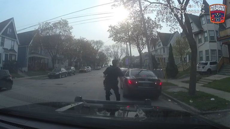 Police officer approaches parked car on residential street.