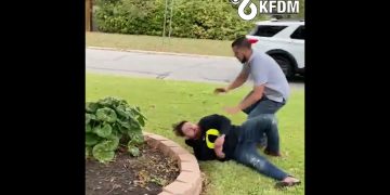 Two people scuffling on grass near road.