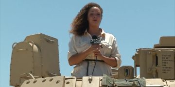 Reporter on military vehicle with microphone.