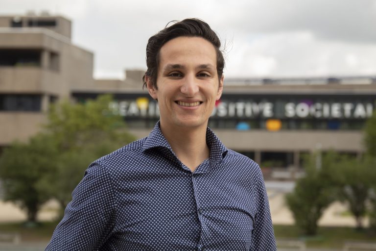 Person smiling outdoors with blurred building background.