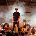 Man with sunglasses surrounded by drums and fog.