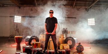 Man with sunglasses surrounded by drums and fog.