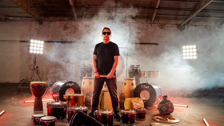 Man with sunglasses surrounded by drums and fog.