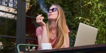 Woman smoking outside with laptop and coffee.