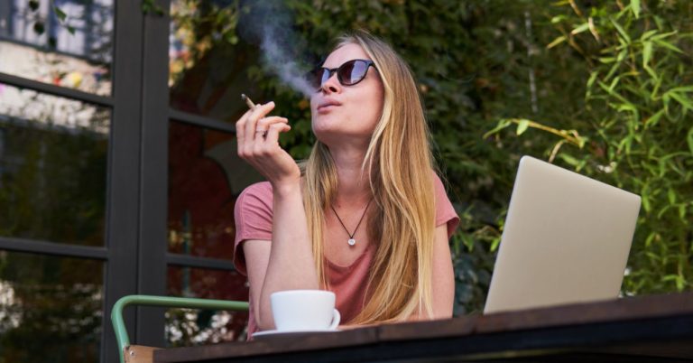 Woman smoking outside with laptop and coffee.