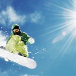 Snowboarder jumps under bright sun with leaf symbol.