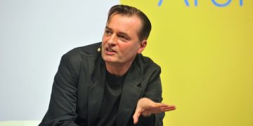 Person speaking at a conference, yellow background.