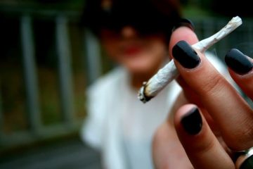 Hand holding a small rolled cigarette outdoors.